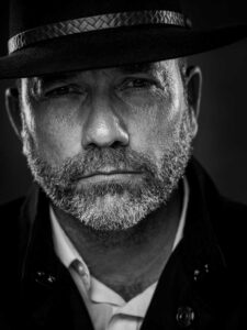 Stoere zwartwit portretfoto van een man met cowboyhoed en baard.
