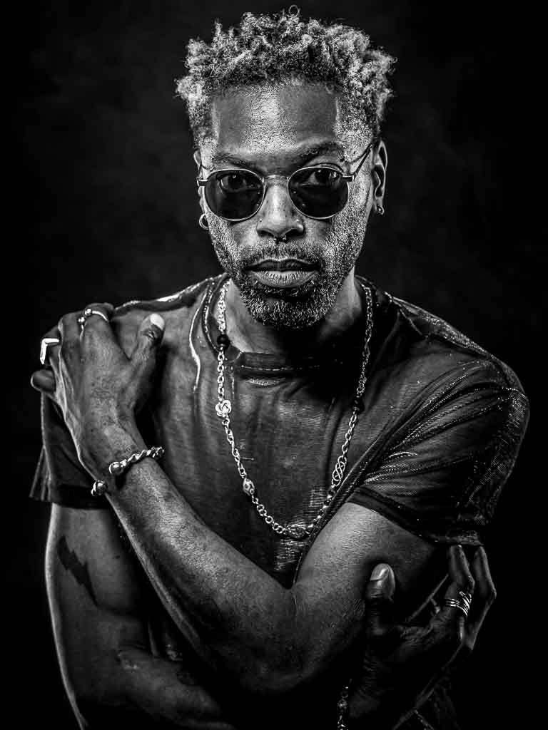 Stoere zwartwit portretfoto van een man met zonnebril en ketting. Gemaakt met flitslicht in min studio in Woerden.